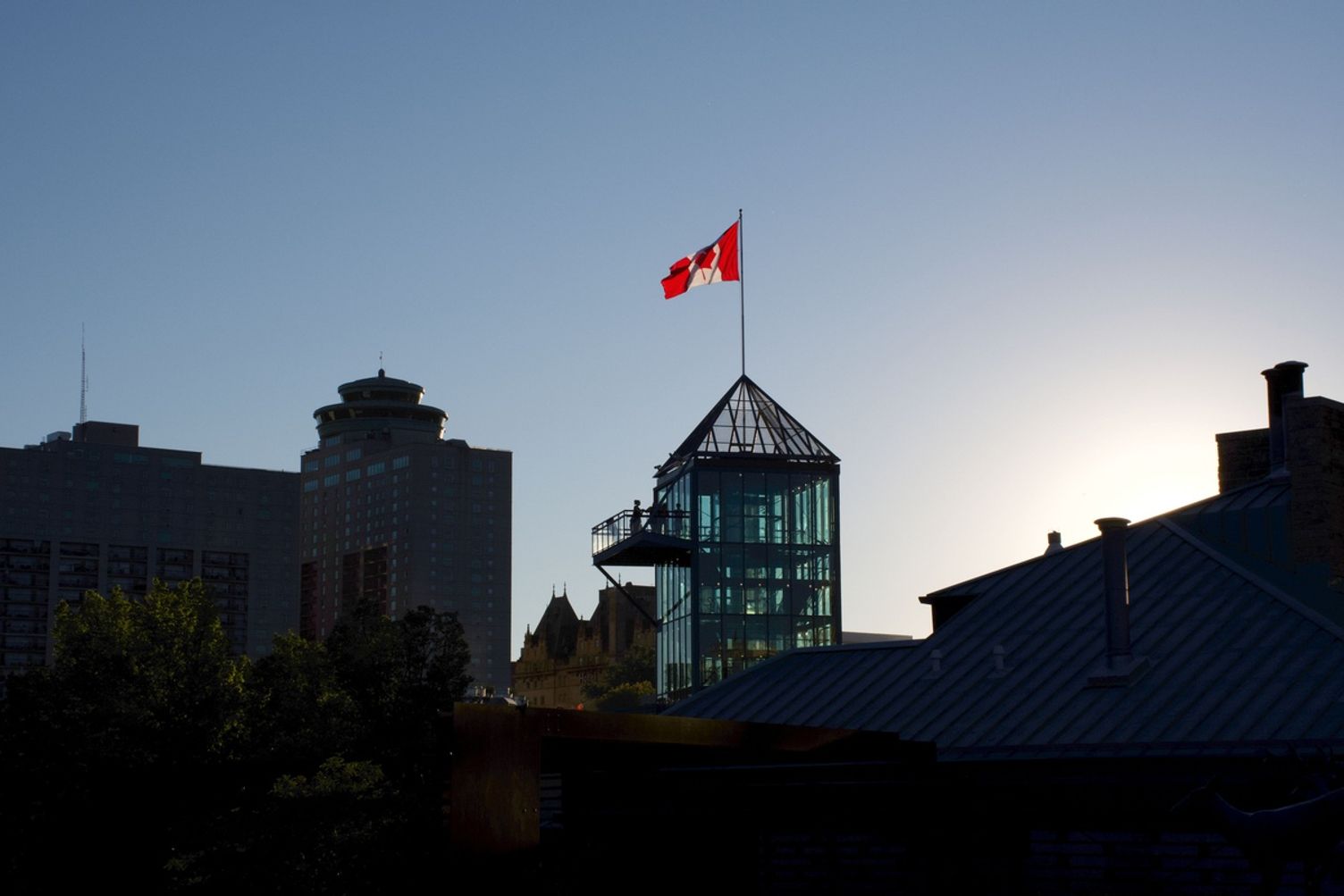 A Canadian flag flying atop a modern glass tower against a clear sky in a city skyline.