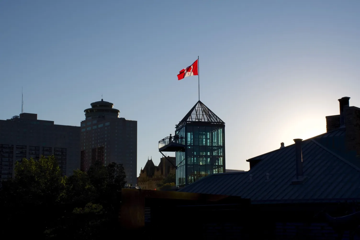 A Canadian flag flying atop a modern glass tower against a clear sky in a city skyline.
