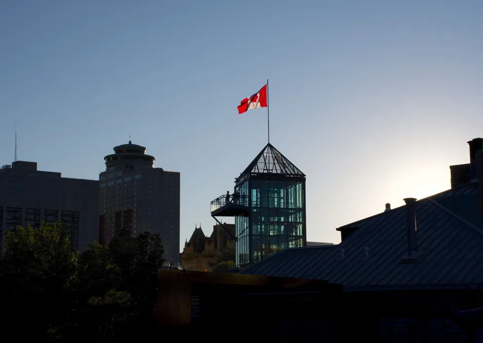 A Canadian flag flying atop a modern glass tower against a clear sky in a city skyline.