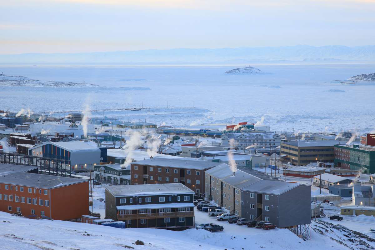 A snow-covered town in Nunavut with smoke rising from buildings beside a frozen bay.