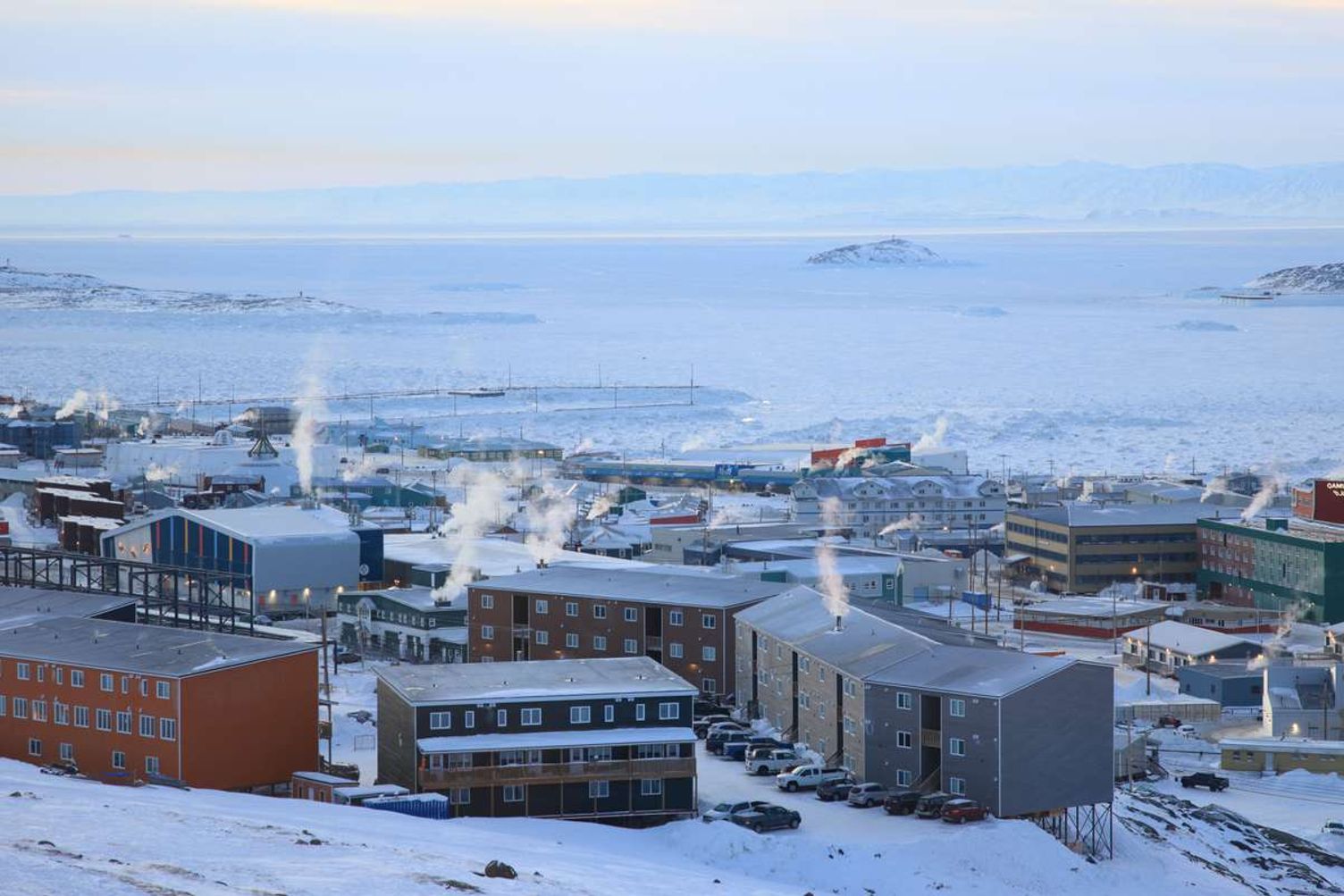 A snow-covered town in Nunavut with smoke rising from buildings beside a frozen bay.