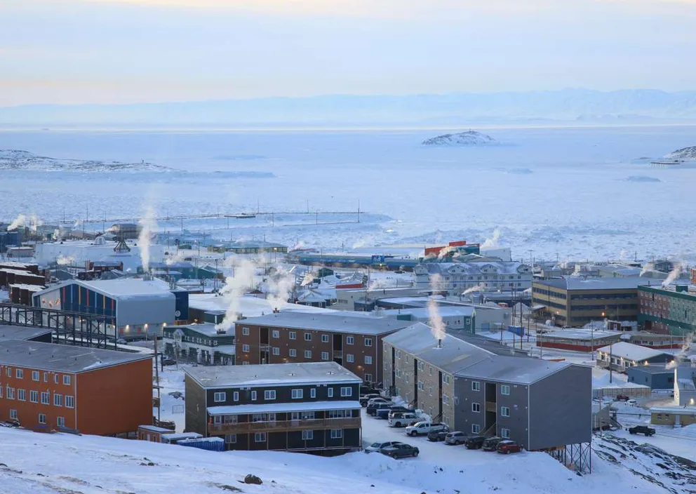A snow-covered town in Nunavut with smoke rising from buildings beside a frozen bay.