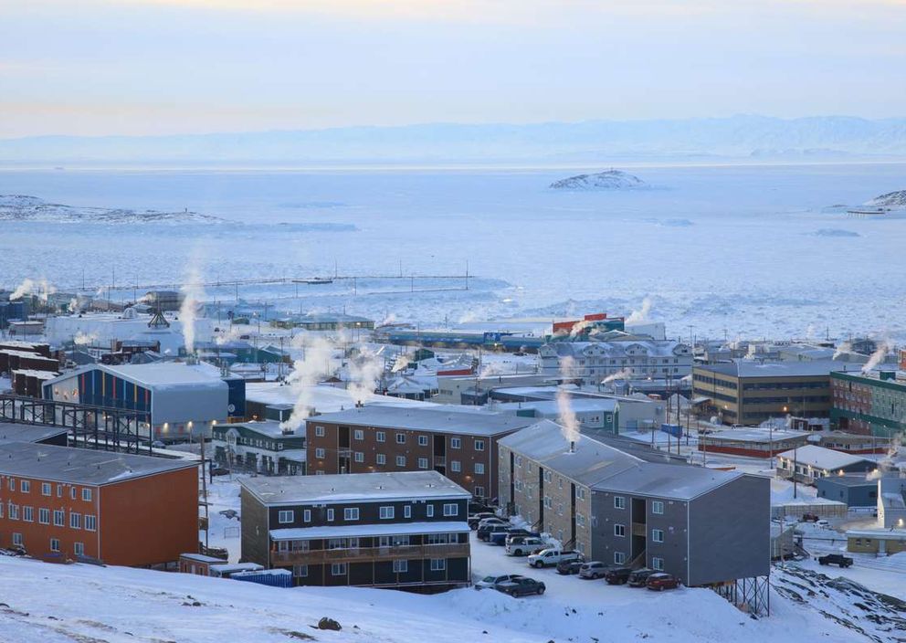 A snow-covered town in Nunavut with smoke rising from buildings beside a frozen bay.