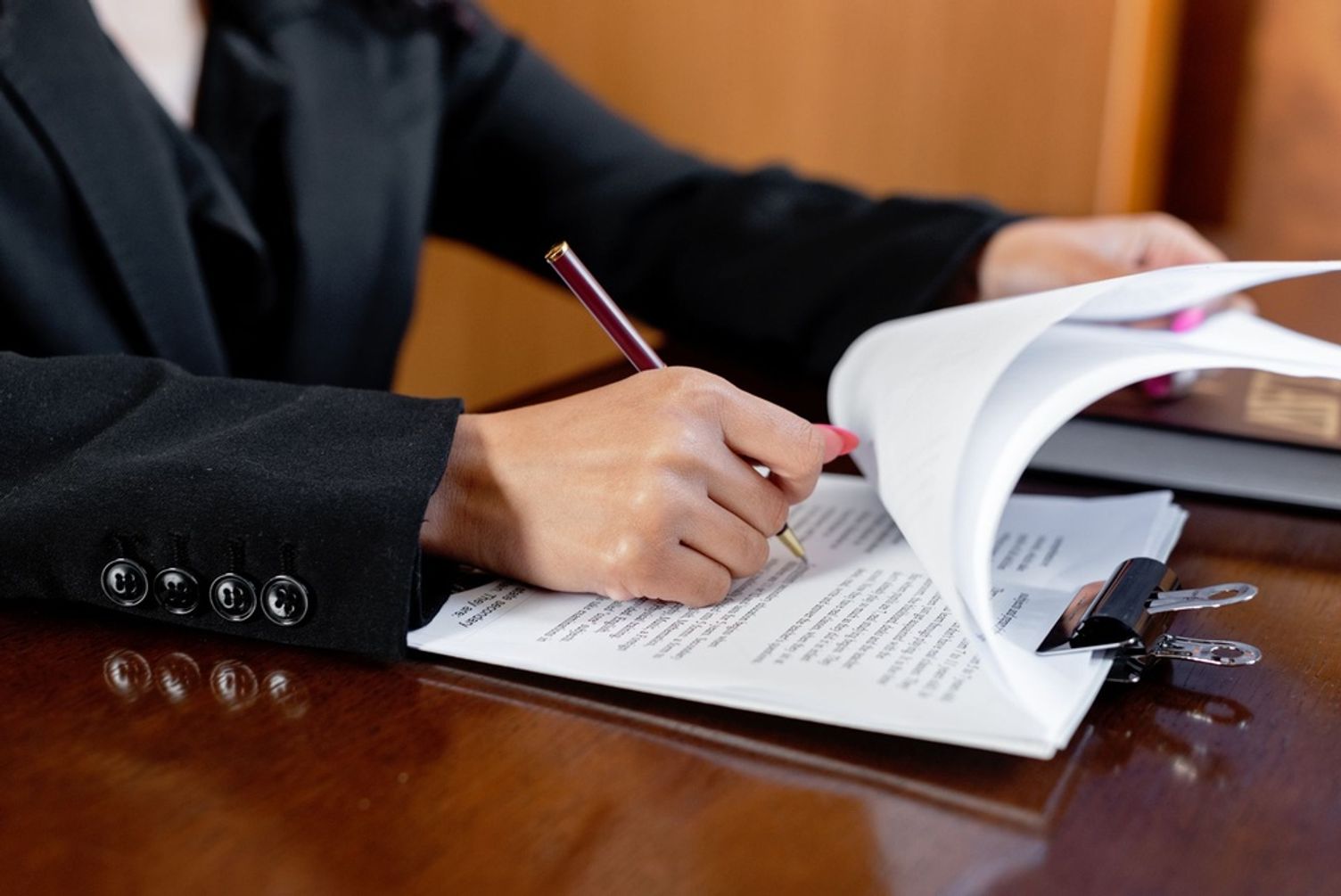 Close-up of a person in a black suit reviewing and signing documents on a clipboard at a desk.