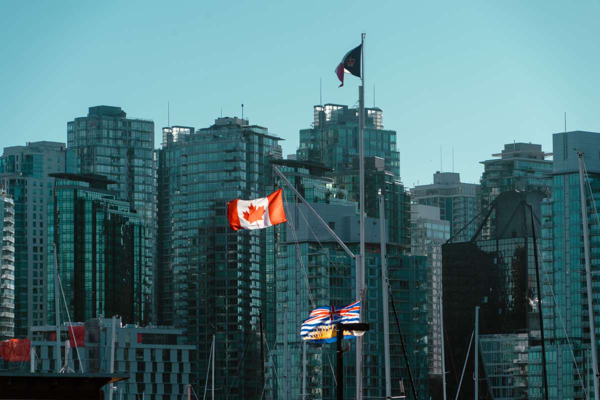 Canadian and British Columbia flags flying in front of modern glass high-rises in Vancouver.