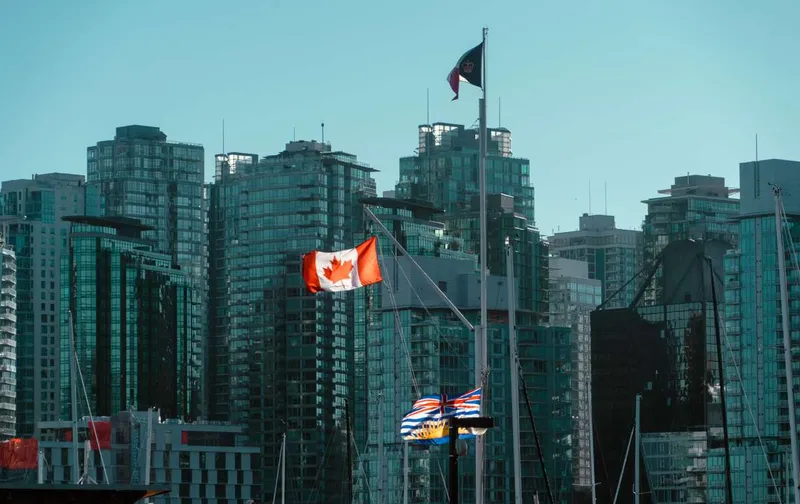 Canadian and British Columbia flags flying in front of modern glass high-rises in Vancouver.