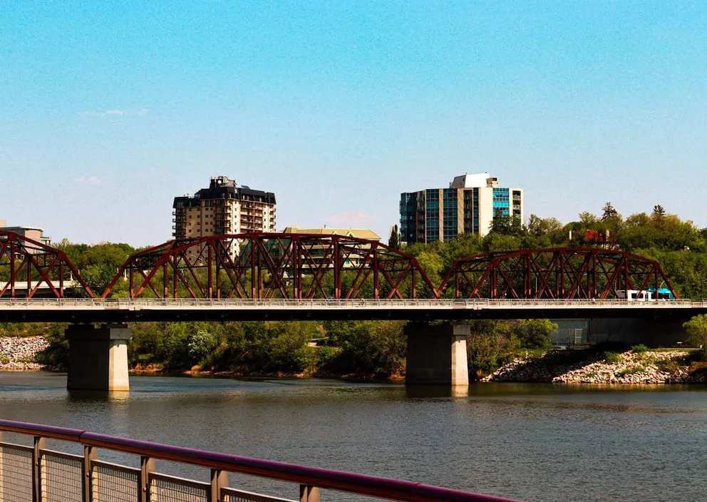 A red steel truss bridge crossing a river with city buildings and trees in the background under a blue sky.