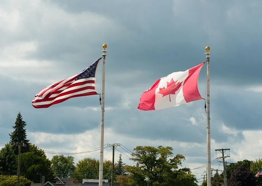 American and Canadian flags flying side by side on flagpoles under a cloudy sky.
