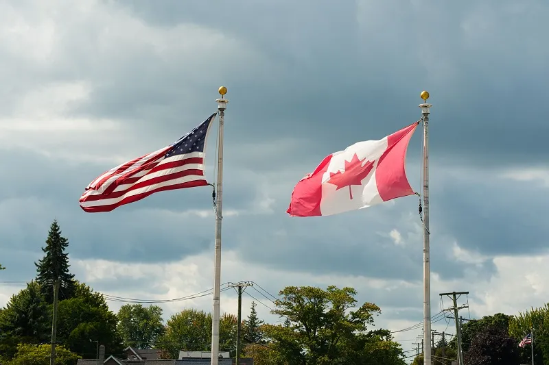 American and Canadian flags flying side by side on flagpoles under a cloudy sky.