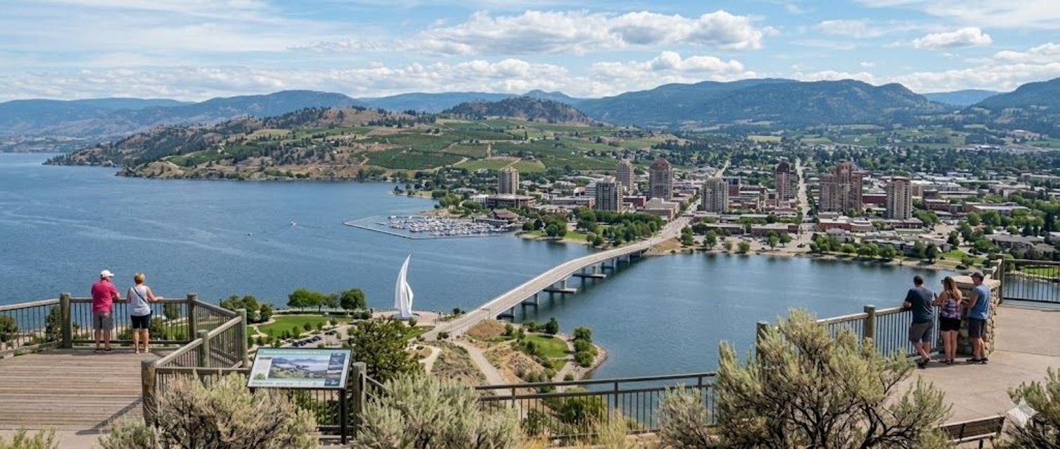 Scenic overlook of Kelowna, British Columbia, showing Okanagan Lake, a bridge, and surrounding hills.