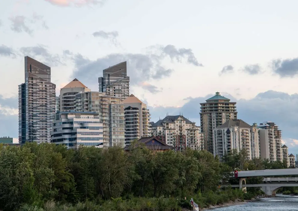 Modern city skyline with tall high-rise buildings rising behind green trees along a riverbank.