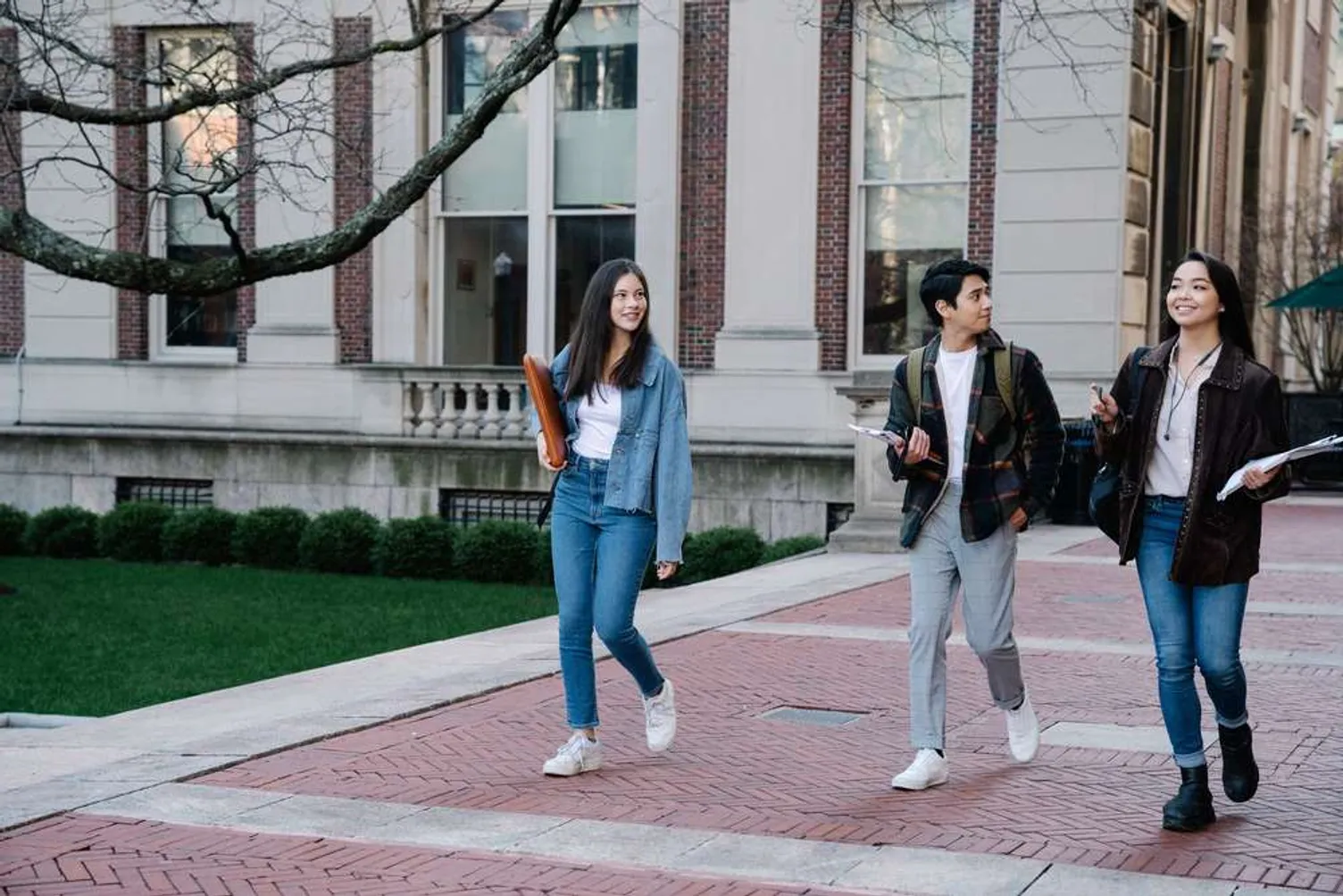 Three university students walk and chat on a brick pathway outside an academic building.