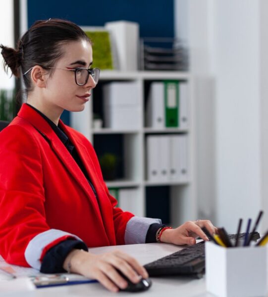 immgration-to-canada (4) Businesswoman in red blazer working on a desktop computer in a modern office.