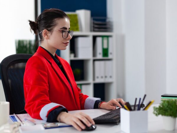 Businesswoman in red blazer working on a desktop computer in a modern office.