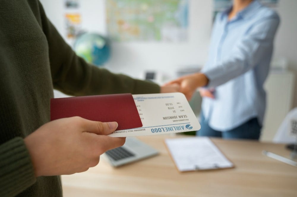 Hand holding a boarding pass and passport, with travel-related items on a desk.