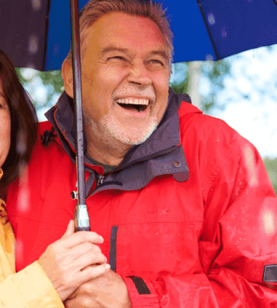 Smiling older couple under umbrella in rain, showcasing happiness and support, relevant to Canadian immigration services.