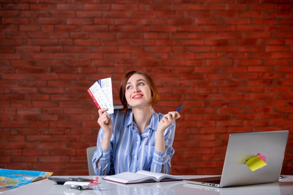 Person holding three airline tickets and a pen at a desk with a map, calculator, notebook, and laptop, preparing for travel.