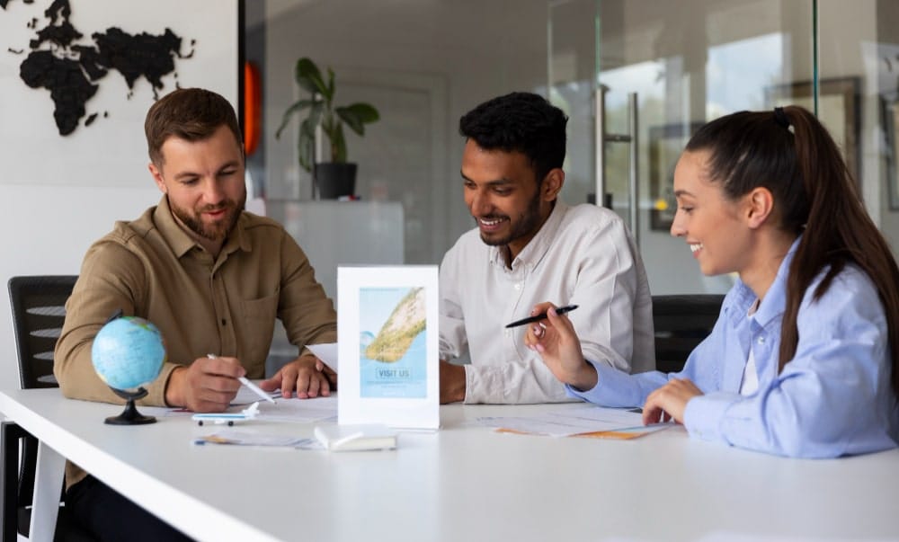 **ALT text:** Three young professionals collaborating at an office table with travel materials.