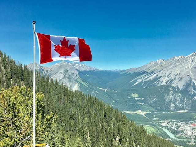 Canadian flag waving on a mountaintop with a panoramic view of forested valleys and rocky mountain peaks under a clear blue sky.
