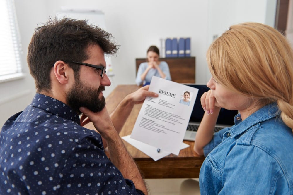 Two people reviewing a resume during a job interview, with a candidate sitting in the background