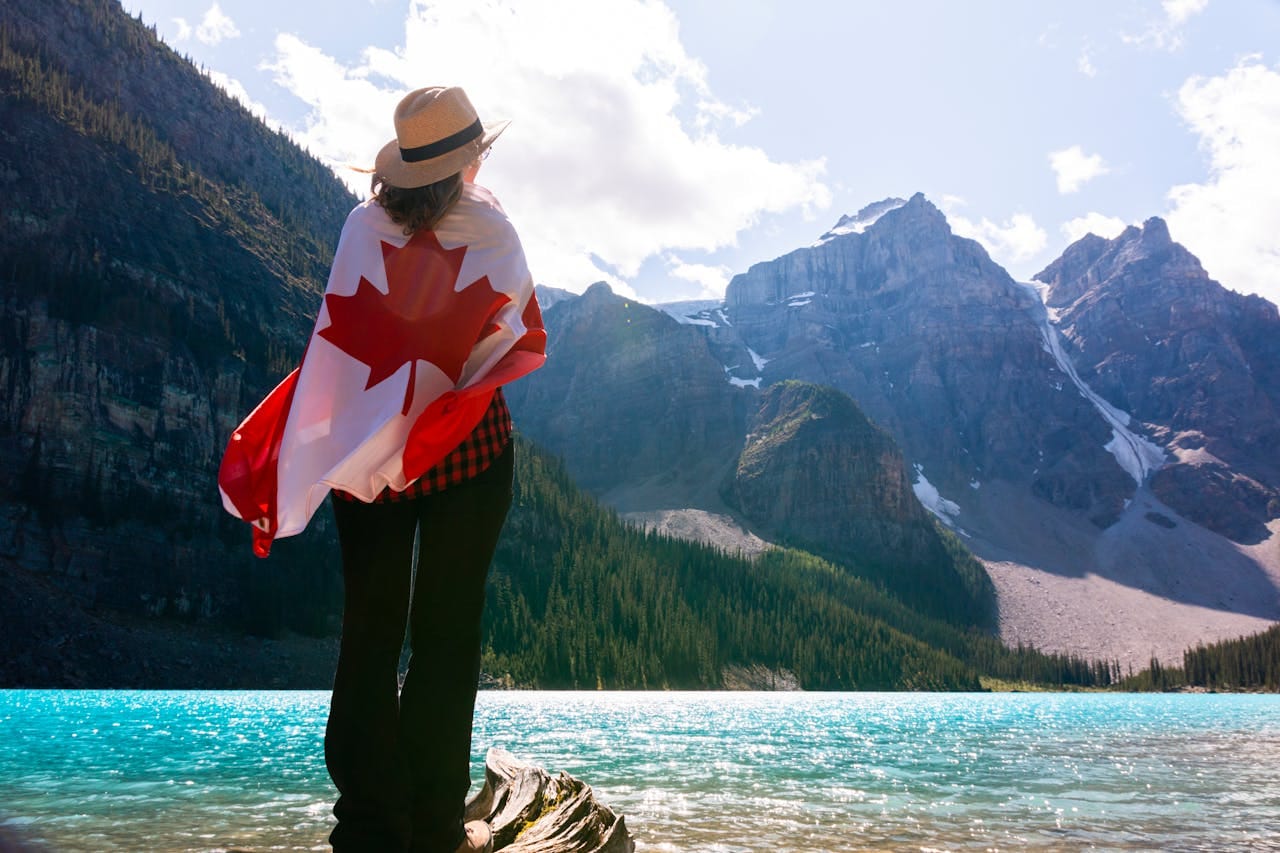 Person wrapped in a Canadian flag looking at a mountain landscape by a turquoise lake.