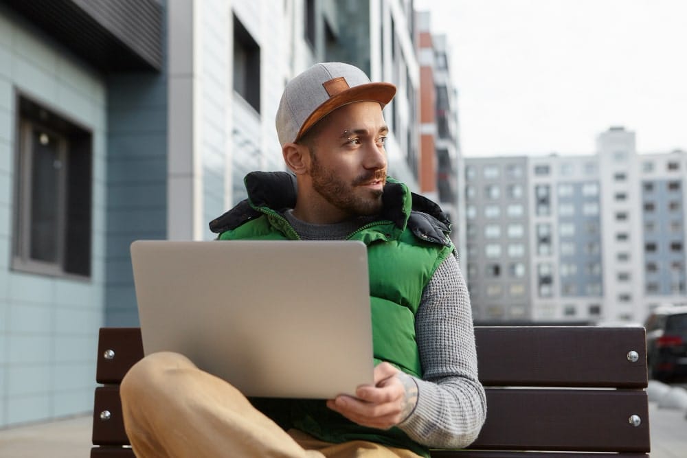Man sitting on a bench outdoors, using a laptop, with modern apartment buildings in the background.