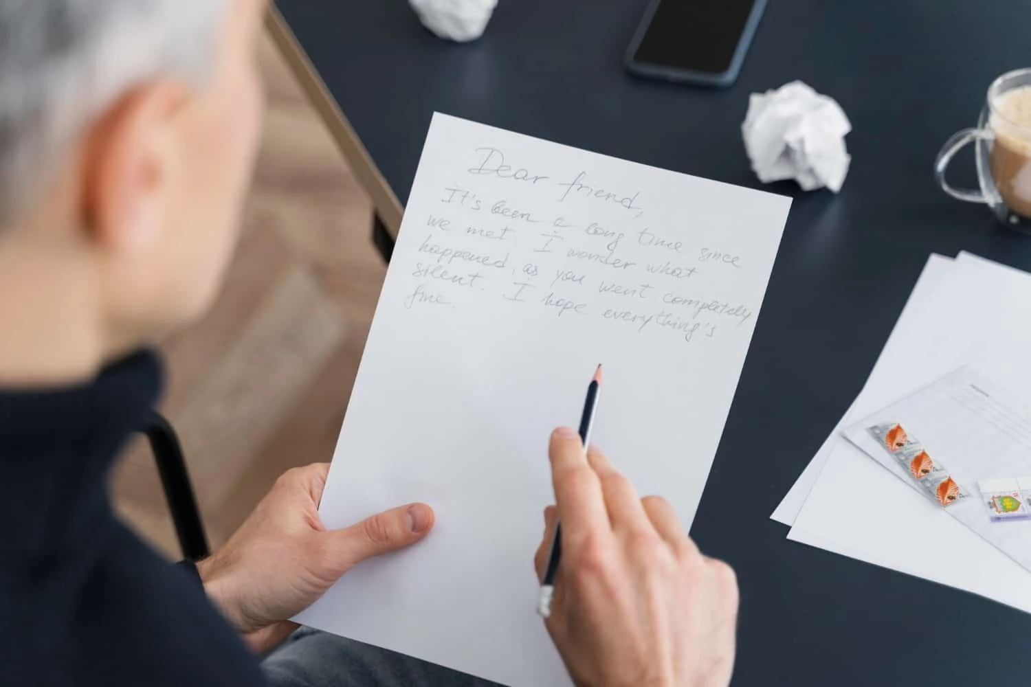 A person holding a handwritten letter that begins with “Dear friend,” while sitting at a desk with crumpled paper, a coffee cup, and postage stamps.
