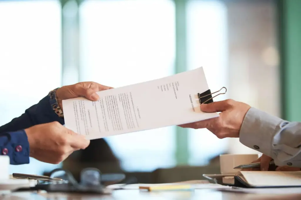 One person handing a stack of official documents clipped together to another person across a desk in a professional setting.