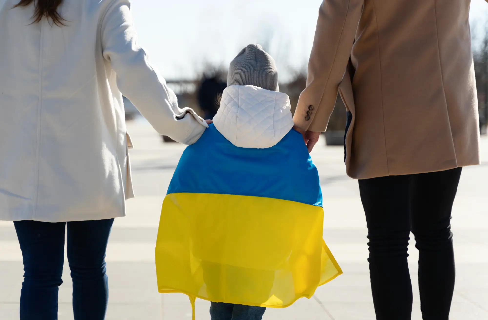Back view of a child draped in the Ukrainian flag, holding hands with two adults, symbolizing support for Ukrainian refugees.