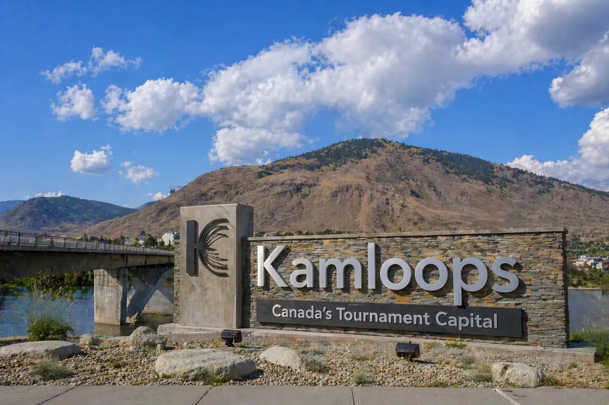 “Scenic riverfront in Kamloops with city landmark sign, bridge, and rolling hills under bright sky”