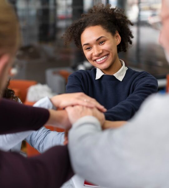 A group of diverse professionals, including a smiling young Black woman with curly hair, placing their hands together in a stack over a table, symbolizing teamwork, collaboration, and unity