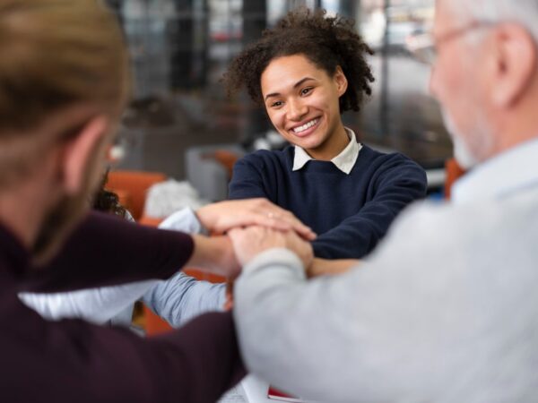 A group of diverse professionals, including a smiling young Black woman with curly hair, placing their hands together in a stack over a table, symbolizing teamwork, collaboration, and unity